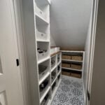 Finished under-stairs closet makeover with white shelving, wood countertops, storage bins, woven baskets, and patterned peel-and-stick floor tile.