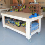 Finished DIY rolling workbench with a large plywood top, bottom storage shelf, casters, and woodworking tools in a garage workshop.