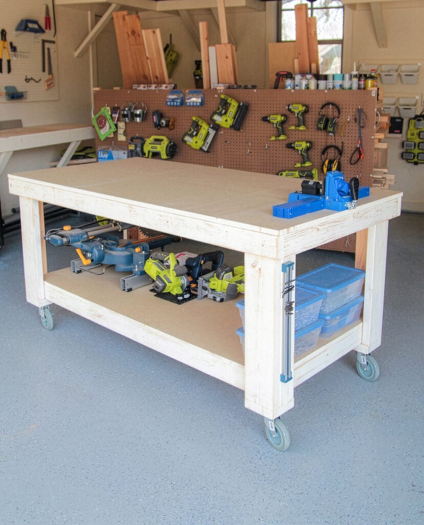 Finished DIY rolling workbench with a large plywood top, bottom storage shelf, casters, and woodworking tools in a garage workshop.